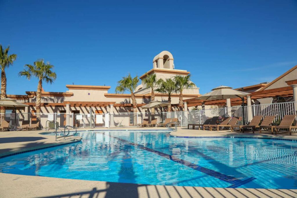 The outdoor swimming pool with lounge chairs and tables under umbrellas awaits guests beneath a clear blue sky. Palm trees sway nearby, while a charming building with a bell tower hints at something special, as the Cavco experience is coming soon.