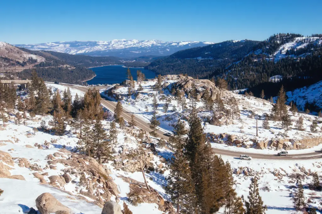 Amid the Winter Wonders, a snowy landscape unfolds with a winding road through bare trees and rocky terrain. A large lake mirrors the distant, snow-covered mountains beneath a clear blue sky, offering one of the perfect Snowy Escapes for those seeking serene beauty.