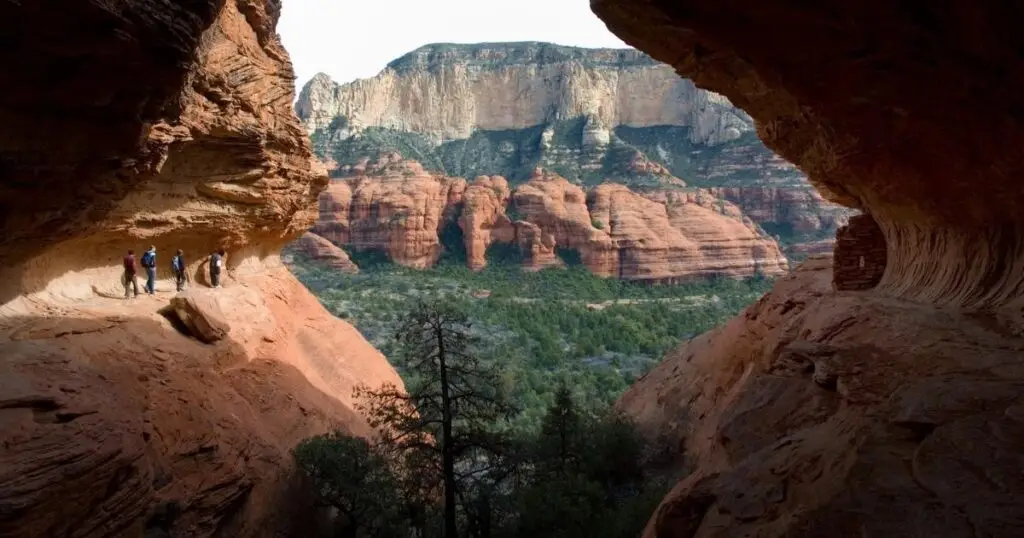 People hiking along a narrow trail through Arizona's regional parks, winding through a large rocky canyon with green vegetation and layered cliffs in the distance.