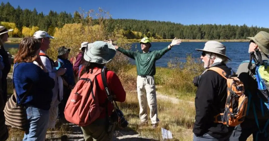 A man in a green shirt gestures while speaking to a group of people near a lake in California, surrounded by seasonal foliage and trees under a clear blue sky, creating an atmosphere of fun and relaxation.