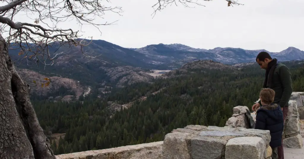 A person and a child stand by a stone wall, gazing over California's forested mountain landscape under a cloudy sky, ready to embark on their grand adventure.