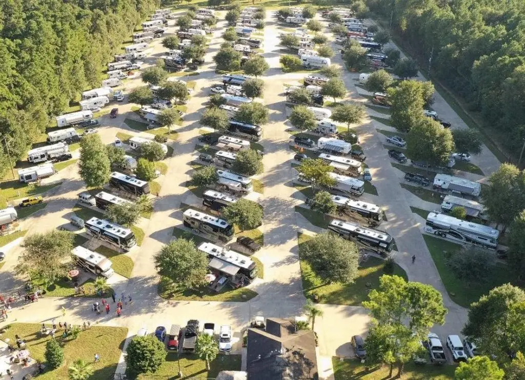Aerial view of Rayford Crossing RV Resort, a large RV park with rows of recreational vehicles and trees, surrounded by greenery. The expanding site shows people near some vehicles.