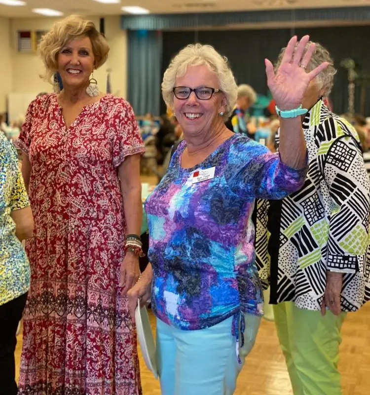 In a vibrant room at Sunrise RV Resort in Apache Junction, Arizona, two women dressed in colorful outfits share a joyful moment as one waves and they both smile warmly amidst the lively crowd.