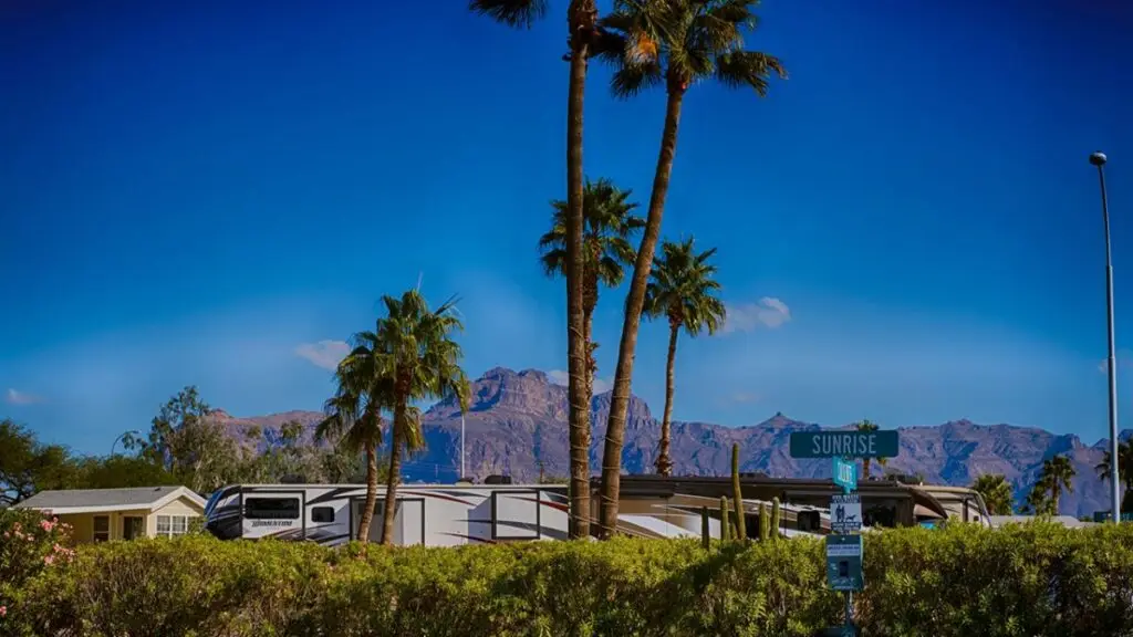 Palm trees and RVs stand against a mountain backdrop under a clear blue sky at Sunrise RV Resort. A street sign reads "Sunrise," capturing the essence of an Arizona retirement dream.
