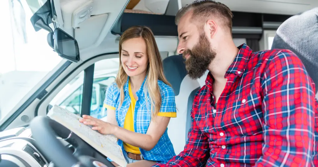 A man and a woman in plaid shirts sit in their RV. The woman, holding a map, is deep into planning their first vacation while the man looks on eagerly.