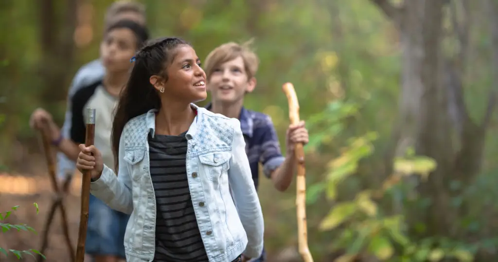 A group of children with walking sticks hike on a wooded trail in Willis, TX, looking up at the trees. It's one of the fun things to do for families exploring nature together.