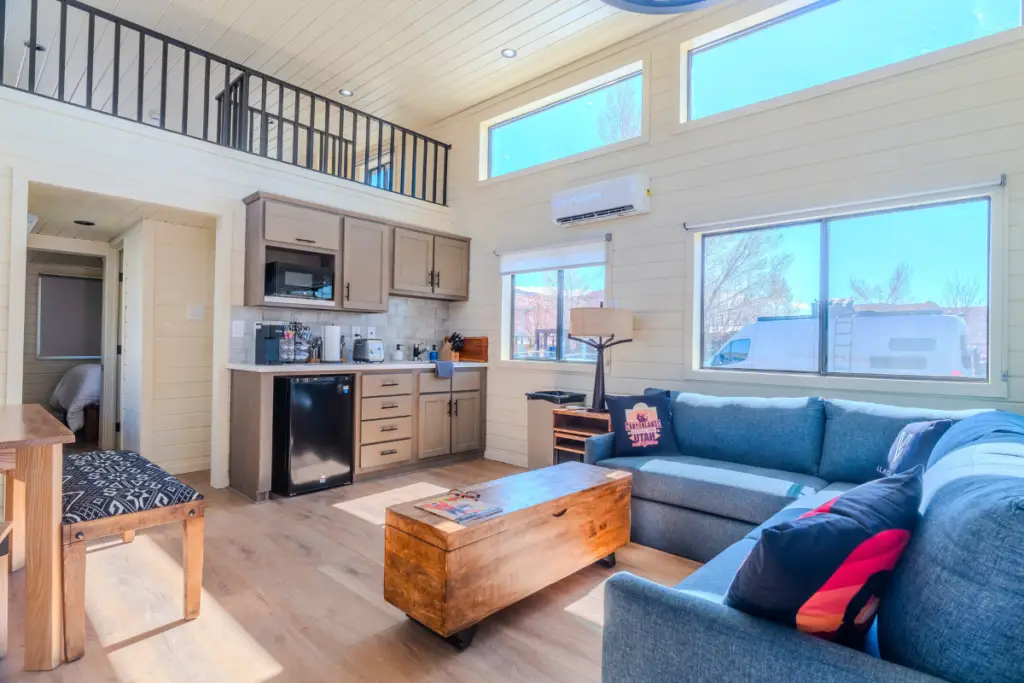 Bright living room with a loft, featuring a blue sectional sofa, wooden coffee table, and kitchenette with appliances. Large windows let in natural light, creating an inviting space reminiscent of the cozy ambiance at Adventure Cabins.