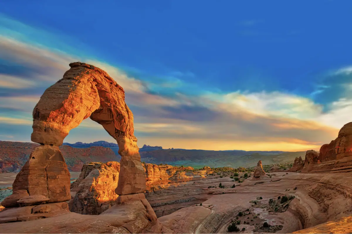 Delicate Arch in Arches National Park, Utah, stands majestically against a vivid blue sky with scattered clouds and a colorful sunset. This rugged terrain offers the perfect backdrop for unforgettable Father's Day adventures amid iconic sandstone formations.