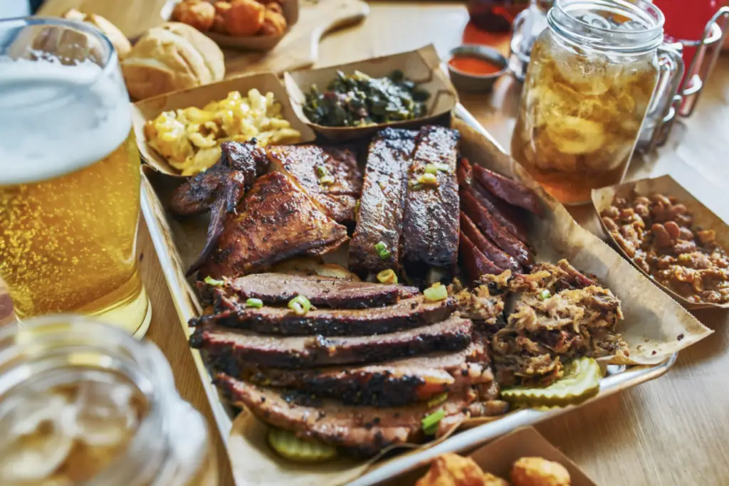 A platter of assorted barbecued meats, including ribs, brisket, and sausage, with sides of pickles, greens, baked beans, and a glass of beer awaits on the wooden table—perfect for a lakeside adventure.