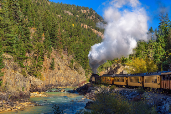A steam train travels through the lush, forested summer landscape of mountain trails alongside a river, with smoke billowing under a clear blue sky. Nearby, the Durango RV Resort offers travelers a perfect retreat amidst nature's beauty.