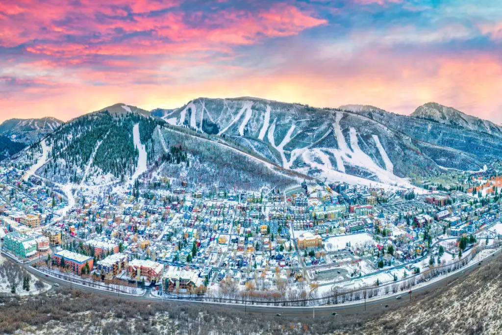Aerial view of Park City, Utah, showcasing a snowy mountainous landscape with a town nestled below. Ski trails weave down the slope, and a colorful sunset sky creates a picturesque backdrop, perfect for New Year's celebrations.