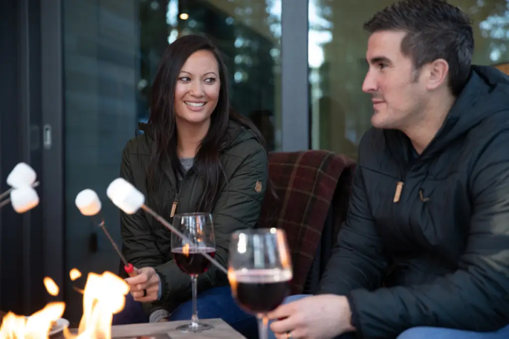 A man and a woman sit by a fire pit at Coachland RV Resort in Truckee-Tahoe, toasting marshmallows and holding glasses of red wine. They appear to be enjoying a casual outdoor Valentine's Day gathering.