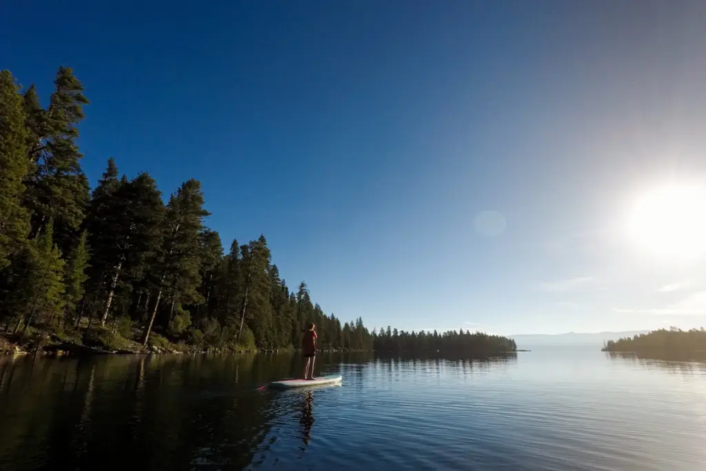 A person paddleboarding on a calm alpine lake near Truckee, surrounded by trees under a clear blue sky and bright early summer sunlight.