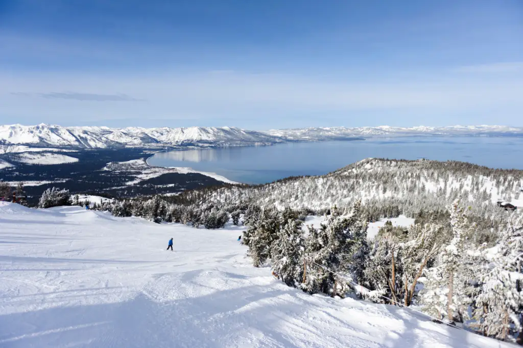 In the picturesque snowy mountain landscape, a skier glides gracefully down the slope, surrounded by trees near Truckee. A large lake and snow-capped mountains stretch into the distance under a clear blue sky, offering perfect end-of-winter activities for those staying at Coachland RV Resort.