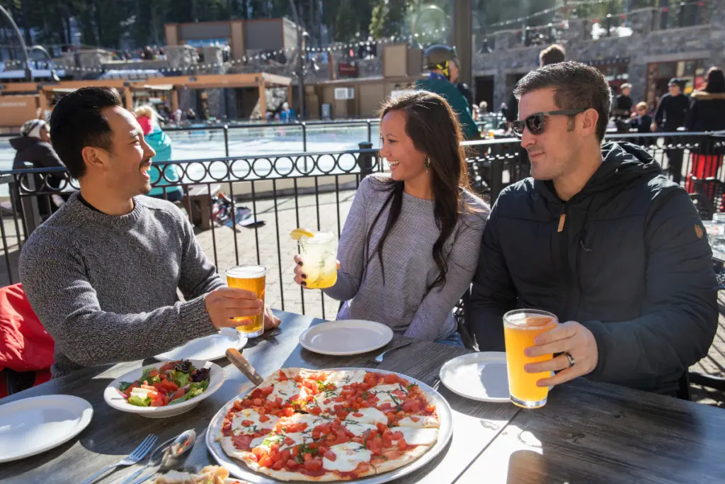 Three people are sitting outdoors at Coachland RV Resort, enjoying drinks and pizza by a pool.