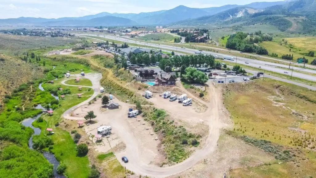 Aerial view of a countryside campground near Park City with several RVs, a winding road, lush greenery, and a stream beside the nearby highway. Perfect for Father's Day adventures, it offers stunning mountains and glimpses of small town buildings in the background.
