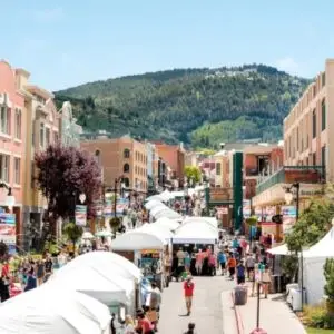 A bustling outdoor street market in Park City features white tents, colorful buildings, and a tree-covered mountain backdrop, perfect for your ultimate summer adventures.