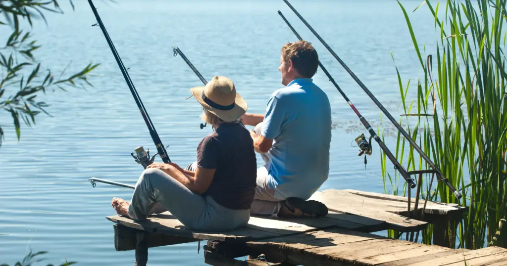 Two people sit on a wooden pier by a calm lake, fishing with rods as tall grass sways around them—a perfect escape among Conroe activities.