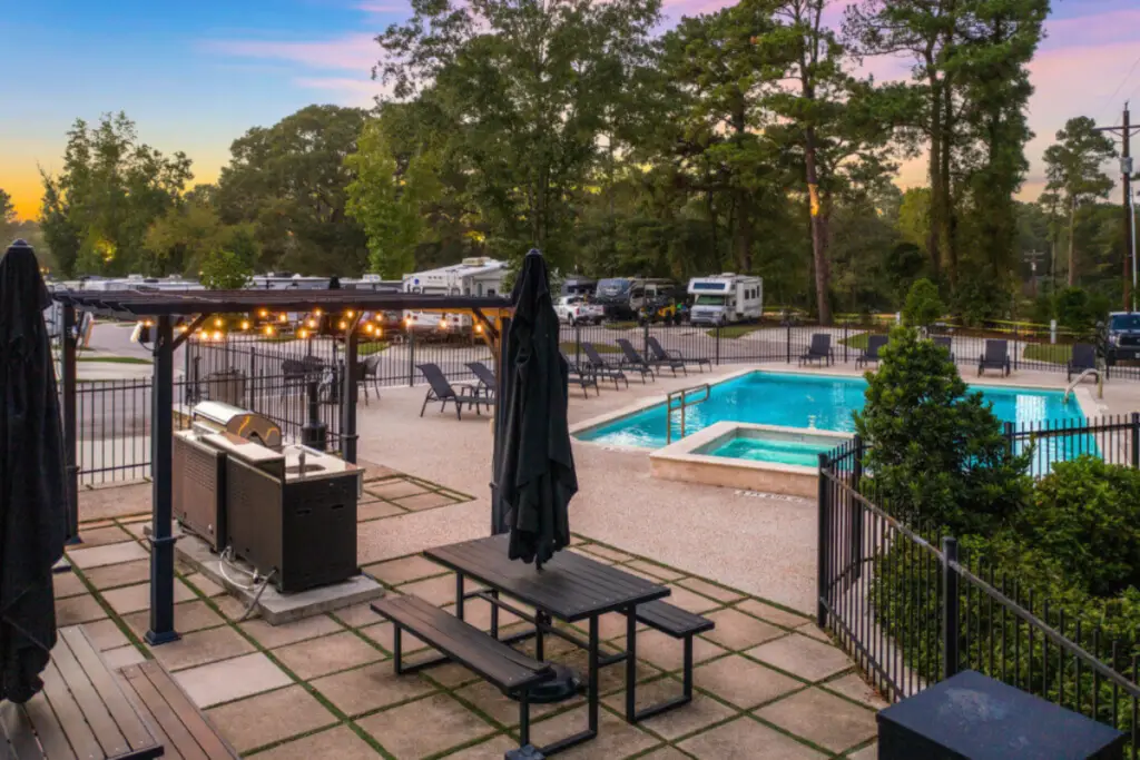 Outdoor area with a swimming pool, barbecue station, and picnic tables. Surrounded by majestic pines, with parked RVs in the background at sunset.