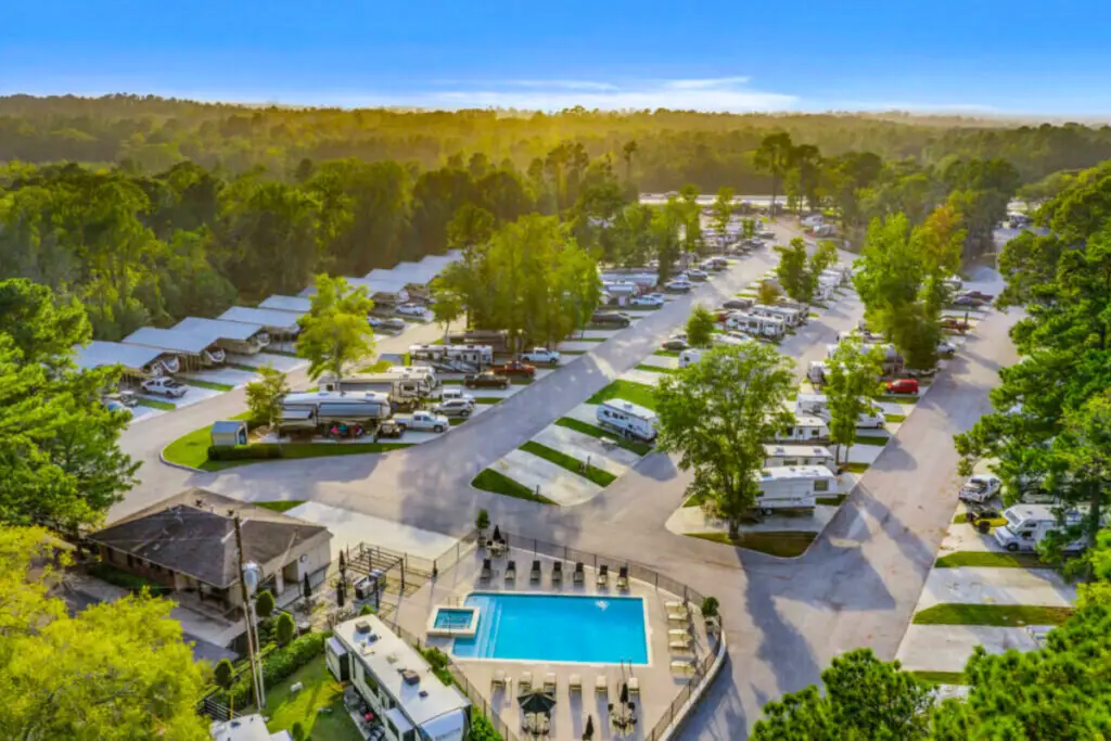 Aerial view of an RV park in a prime location surrounded by trees, featuring parked RVs, cabins, and a rectangular swimming pool with lounge chairs in the foreground, offering visitors the serenity of nature.