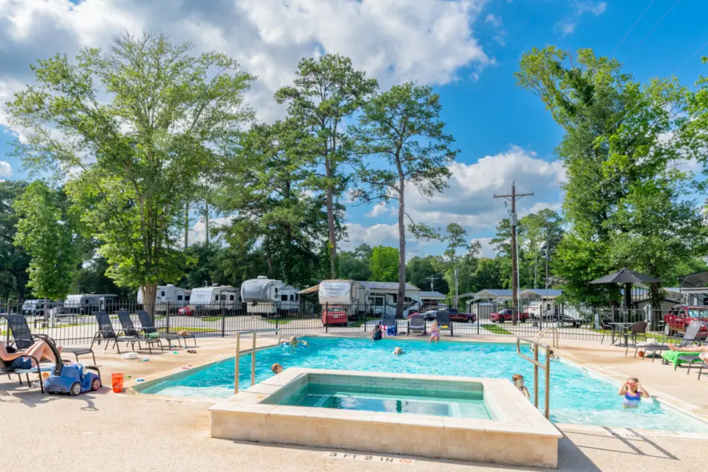 Children are playing joyfully in the outdoor swimming pool, surrounded by lounge chairs and trees at the RV park. Several RVs are parked in the background under a clear blue Texas sky, capturing the essence of leisure near Willis.
