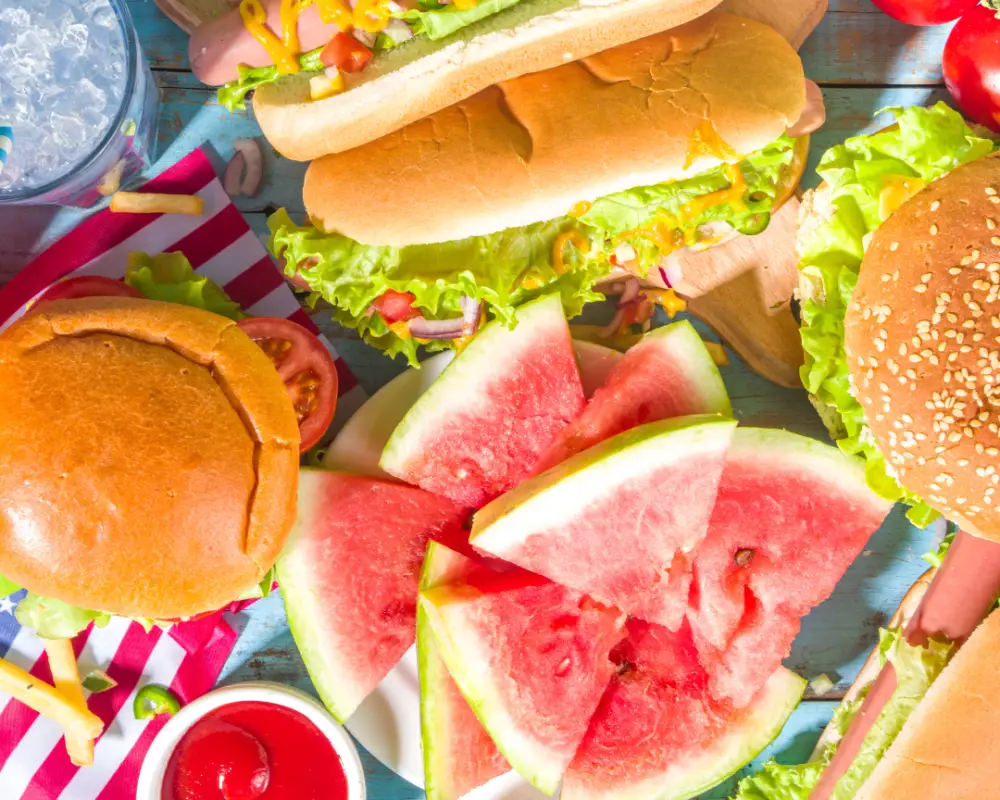 An overhead view captures hot dogs, burgers, and a plate of watermelon slices, evoking the spirit of Memorial Day. There's also a drink, french fries, ketchup, and a tomato on the blue wooden table at Majestic Pines RV Resort.