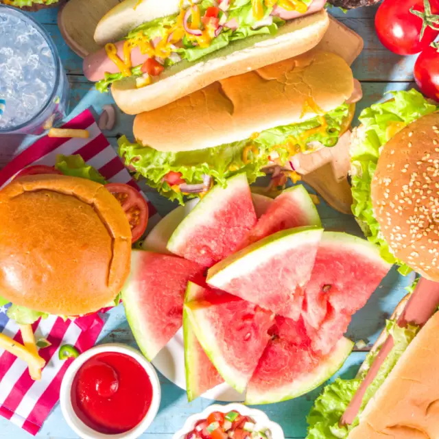 A table at Majestic Pines RV Resort is set for Memorial Day, featuring hot dogs, a burger, sliced watermelon, cherry tomatoes, a refreshing drink with ice, and an array of condiments.