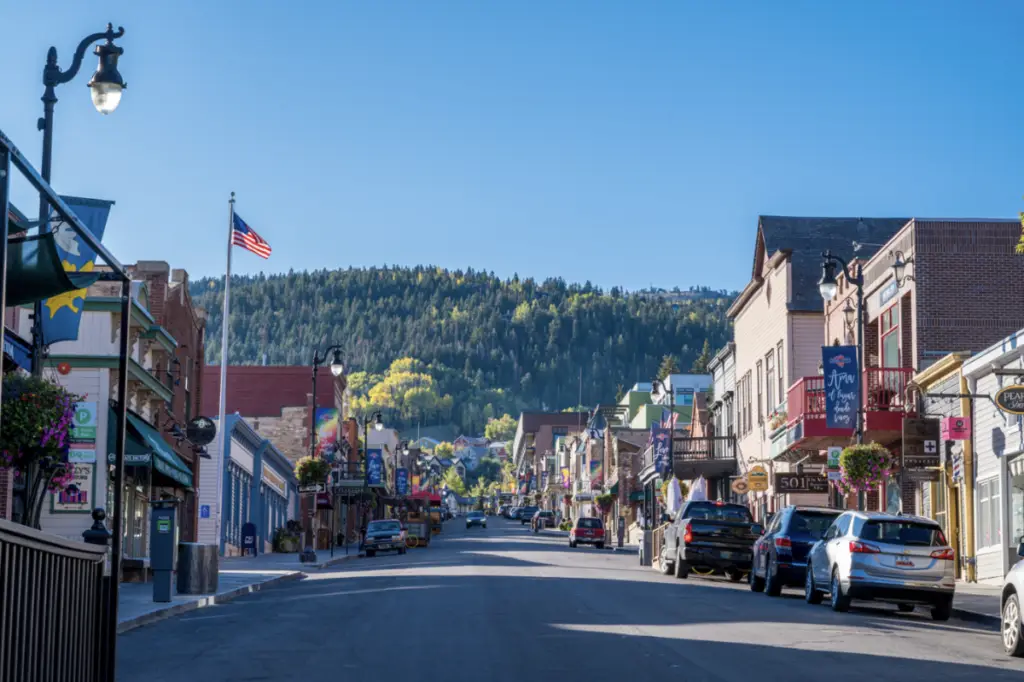 Street view of a small town adorned with an American flag, parked cars, storefronts, and tree-covered hills under a clear blue sky, creating an unforgettable scene reminiscent of a Memorial Day parade.