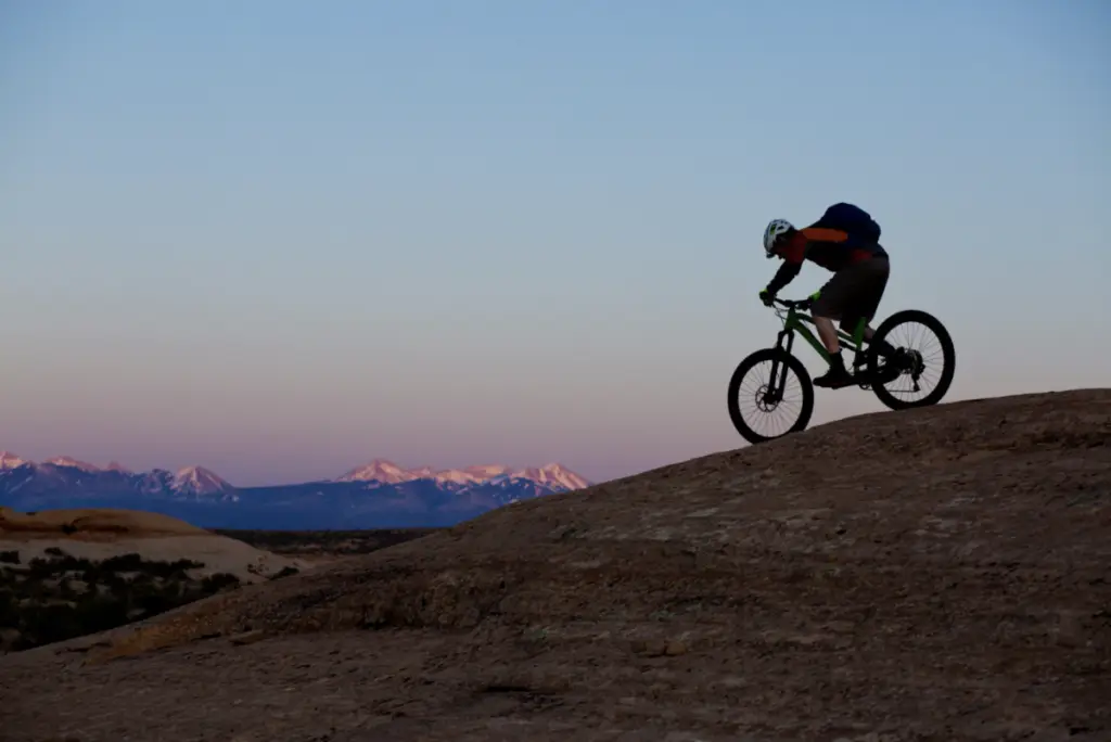 A cyclist wearing a helmet rides uphill on a mountain trail in Moab at sunset. Distant snow-capped peaks are visible under the clear sky, perfectly framing this iconic bike adventure.