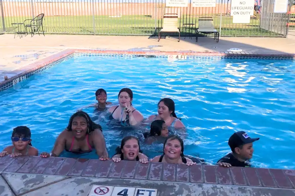 A group of adults and children enjoy a refreshing swim at the Amarillo RV Resort pool, near the edge, with a "No Lifeguard on Duty" sign visible in the background. It's a perfect Memorial Day gathering spot for relaxation and fun.