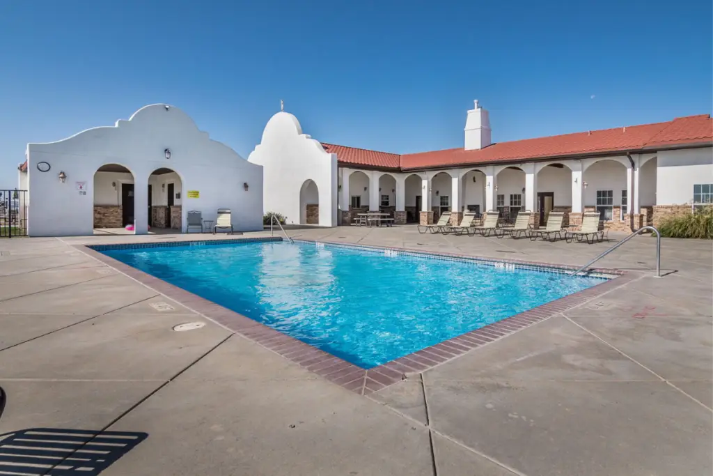 Outdoor pool area with clear blue water, surrounded by a flat concrete deck. White stucco cottages with red tile roofs and arched entrances are in the background under a crystal-clear Amarillo sky.
