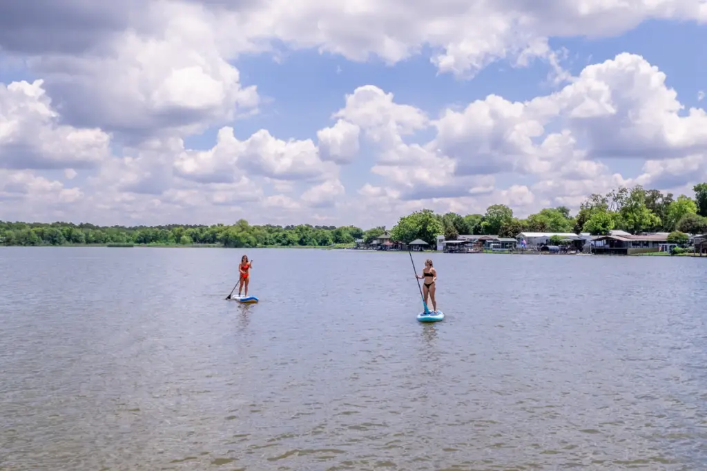 Two people paddleboarding on a calm lake under a partly cloudy sky, enjoying their time at Water's Edge RV Resort, with shoreline trees and houses in the background.