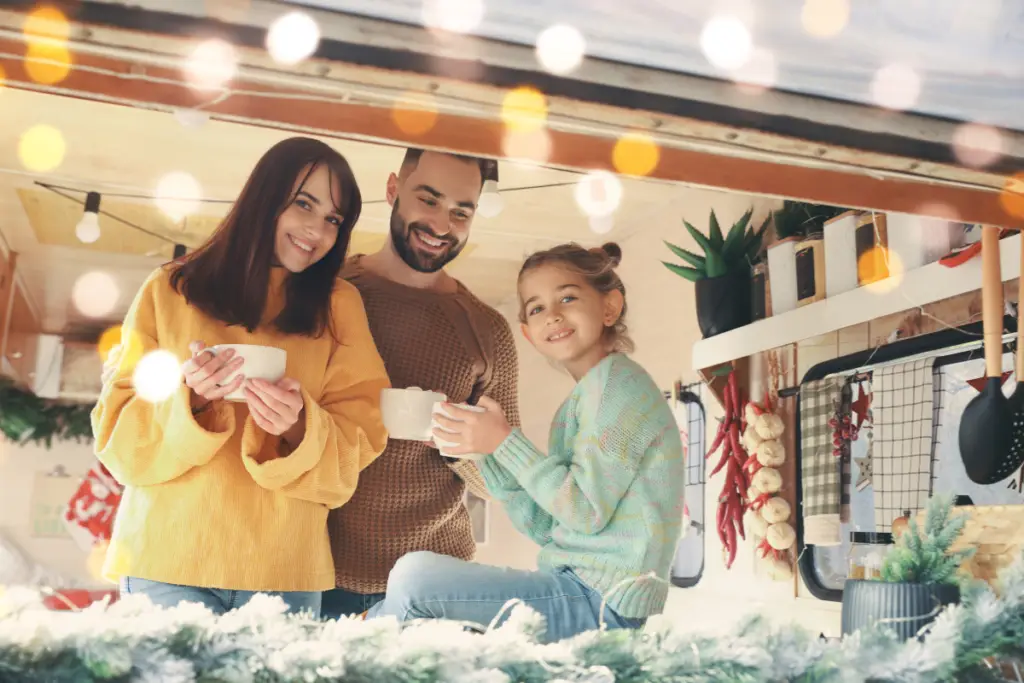 A smiling family of three holding mugs stands in a cozy, decorated kitchen with hanging plants and lights, soaking up the holiday spirit.