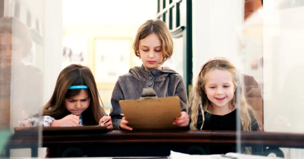 Three children focus on writing as they stand behind a wooden desk, with papers and clipboards in hand, inside a well-lit room at the Children's Museum, making for fun times in The Woodlands.