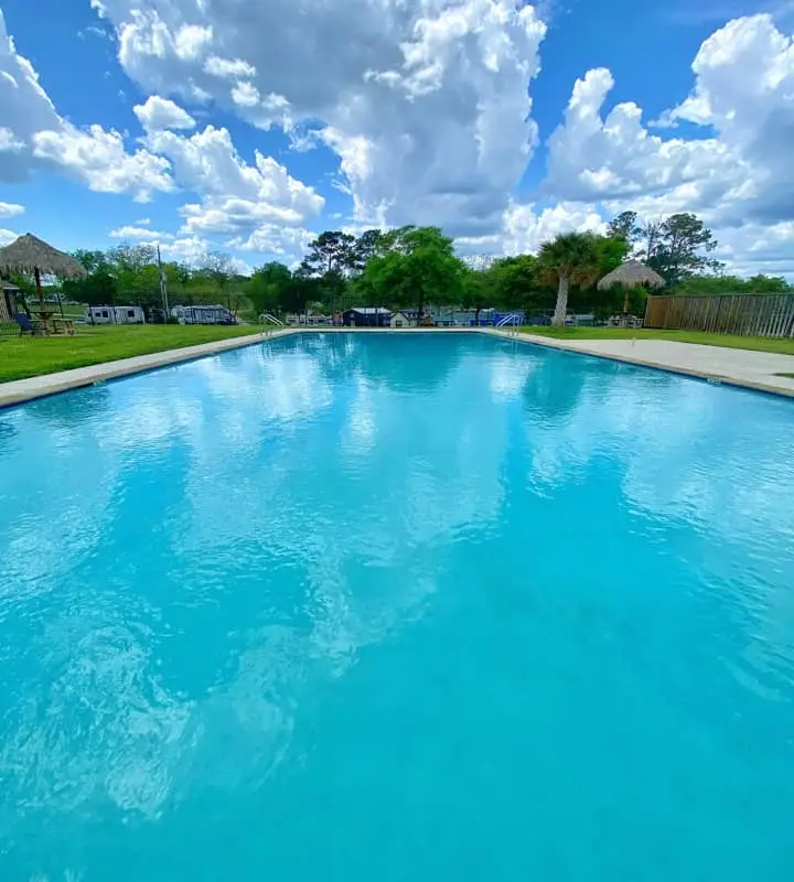 A large outdoor swimming pool sits gracefully at the water's edge, surrounded by lush grass and trees beneath a blue sky with clouds.