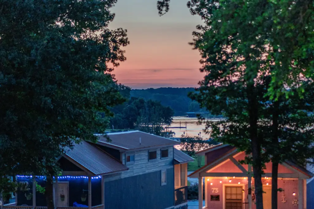 A serene lakeside scene at sunset with two cozy cabins surrounded by trees at Water's Edge RV Resort. The sky is an orange hue, and Lake Conroe reflects the stunning colors of the sunset—perfect for a Spring Break escape.