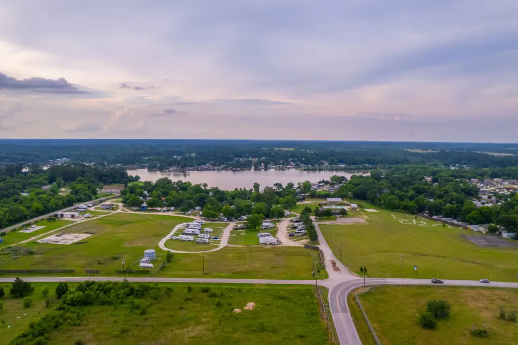 Aerial view of a rural area near Lake Conroe, featuring a road, open fields, scattered buildings, and a small lake with public access surrounded by trees under a cloudy sky.