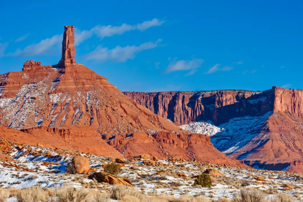 In December, the red rock formations of Moab, Utah stand majestic with their prominent spires under a clear blue sky. Partially dusted in snow, they rise from the rugged desert landscape, creating a mesmerizing and serene winter scene.