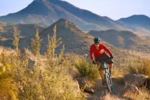 A person wearing a red shirt rides a mountain bike on a trail through a desert landscape with mountains in the background.