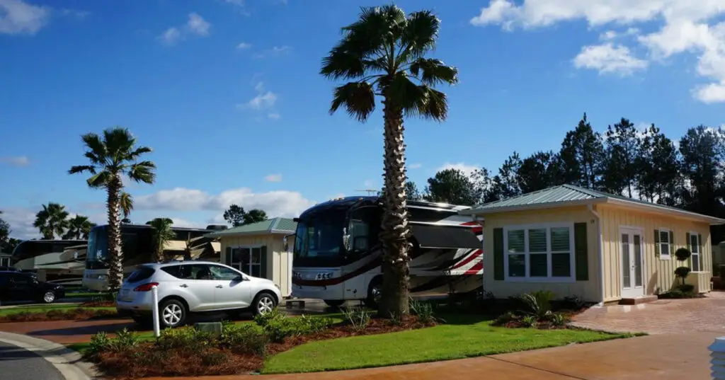 An RV is parked beside a cozy small house and a silver car, with palm trees swaying under the clear blue sky—a perfect scene for a Gulf Shore vacation.