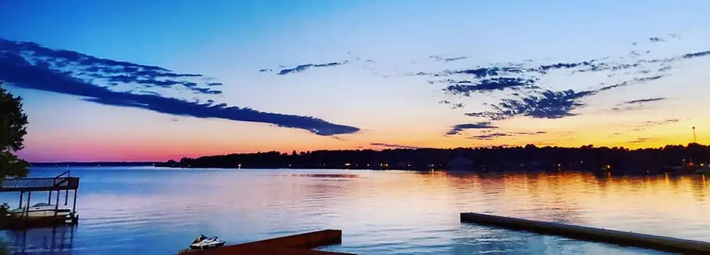 Sunset over Lake Conroe with a dock in the foreground, silhouetted trees along the horizon, and streaked clouds in the summer sky. Book early to capture this serene beauty.