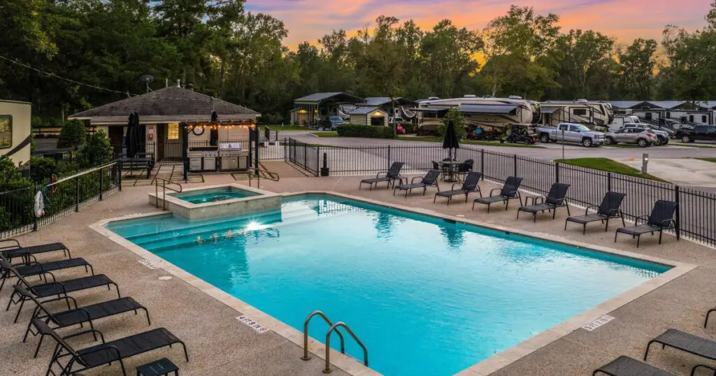 The outdoor swimming pool area, complete with a jacuzzi, is surrounded by lounge chairs and fenced for privacy, offering a luxurious camping experience in an RV park at sunset.