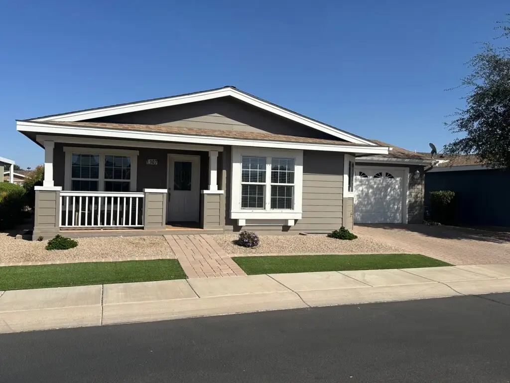 The single-story gray house, reminiscent of the classic F107 design, features a welcoming front porch and elegant white trim. Its two-car garage stands ready beneath a clear blue sky, embodying simplicity and style.