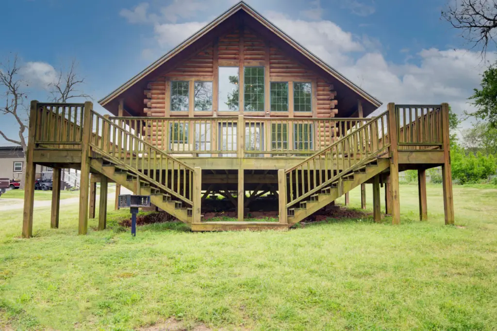 Nestled by Water's Edge, this wooden log cabin features large windows and dual staircases leading to an elevated entrance, all set on a grassy lawn. A perfect retreat for those seeking serenity near Lake Conroe.