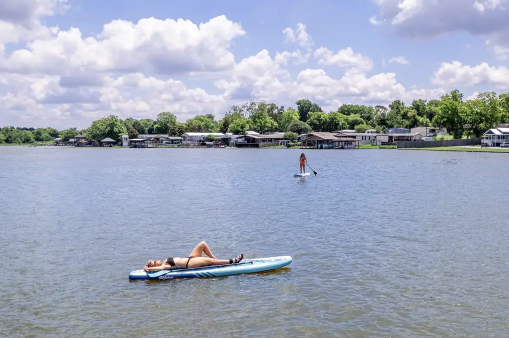 Two people on paddleboards in a serene lake: one laying down basking in the sun, the other standing and paddling. Houses and trees line the distant shore under a partly cloudy sky, perfect for an RV Getaway at Rayford Crossing, where comfort meets nature.