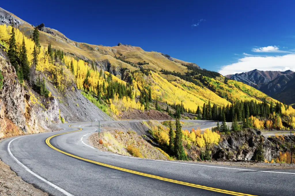 A winding road in Durango curves through a mountainous landscape, offering an outdoor adventure amidst vibrant yellow autumn foliage and coniferous trees under a clear blue sky.