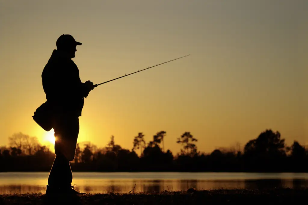 Silhouette of a person fishing at sunrise near the water’s edge of Lake Conroe, surrounded by trees in the background, capturing a moment of pure serenity.