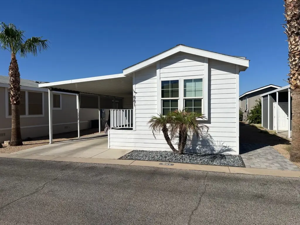 A charming white cottage home sits on Lot 881, featuring a small porch with two palm trees and a concrete driveway under the clear blue sky.