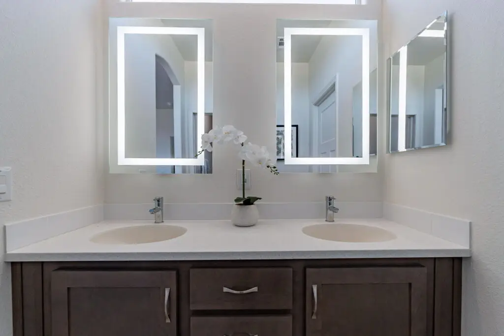 A bathroom vanity with two sinks and a central orchid plant. Two illuminated mirrors and a wall-mounted mirror are above the counter.