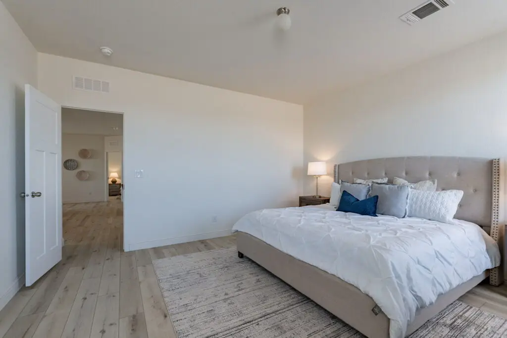 Bedroom with a large bed, tufted headboard, white bedding, blue accent pillows, wooden side table with a lamp, light wood flooring, an open door, and wall decor in view.